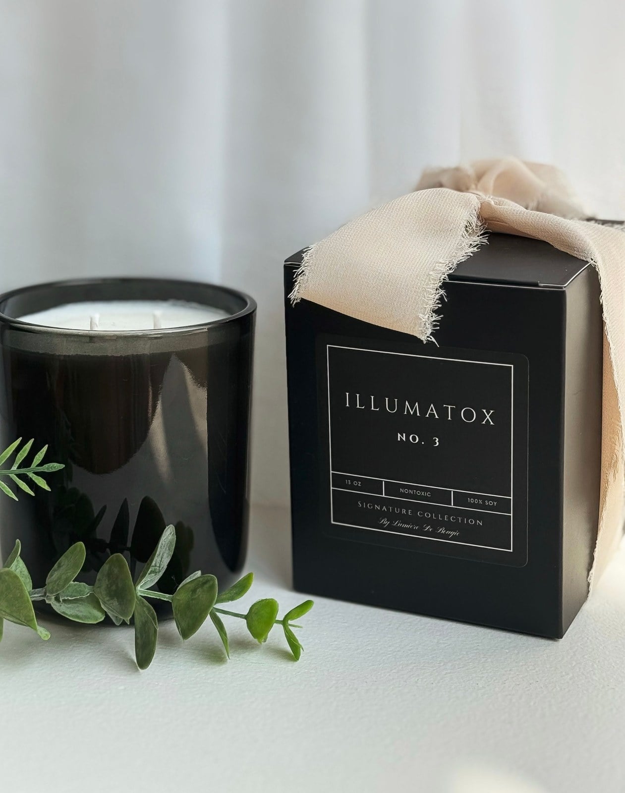 A boxed Illumatox Signature Scents candle with a cream ribbon, photographed beside eucalyptus